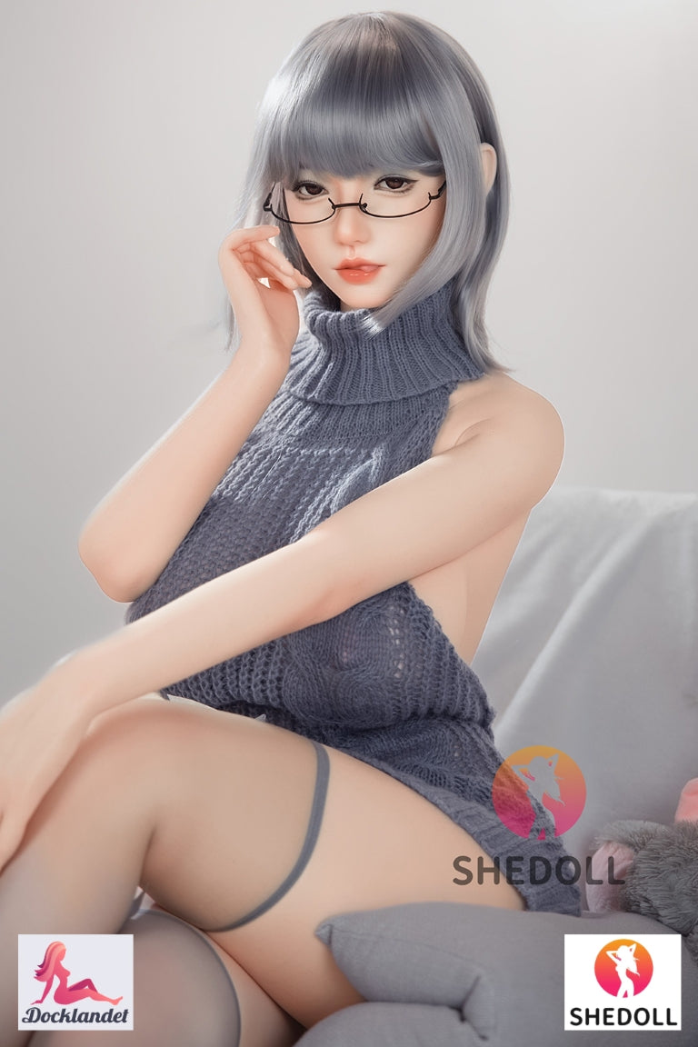 ealistisk Shedoll sexdocka med glasögon och grå polokrage-klänning poserar halvliggande i soffa bredvid mjukt kanin-gosedjur – silikon kärleksdocka hos Docklandet